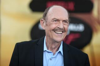 John Ashton in a blue button-up shirt, black blazer smiling against a red and gold backdrop