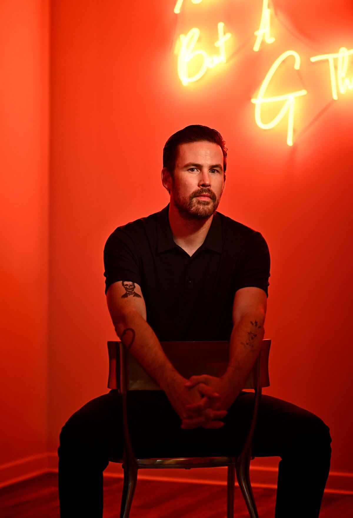 A bearded man in black sits for a portrait in a room bathed in red by neon.
