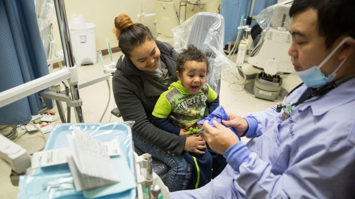 Dentist Gabriel Guevara dons gloves as Angel Grant holds son Josiya, 2, for an oral exam at the Antelope Valley Community Clinic.