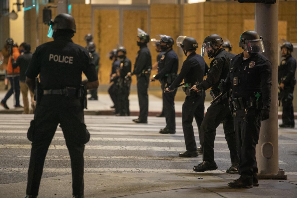 A line of LAPD officers in riot gear stand on a city street.