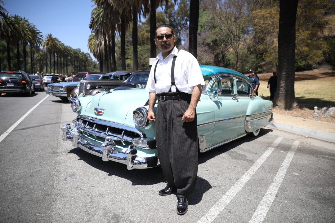 Pedro Haro, in loose pants with pegged ankles and suspenders, stands in front of a classic car.