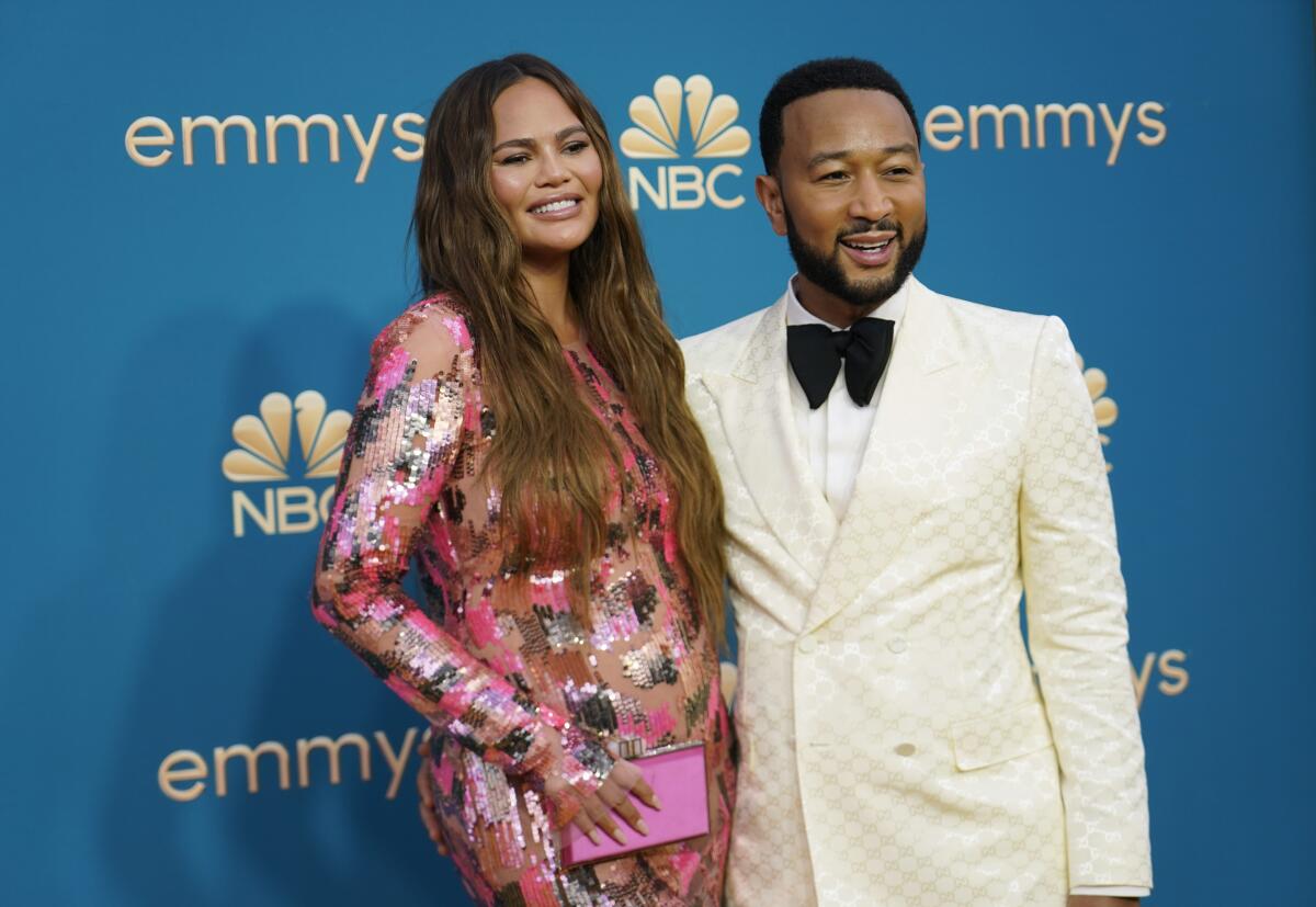 A woman with long brown hair wearing a patterned dress and a man with short black hair and a beard wearing a white suit