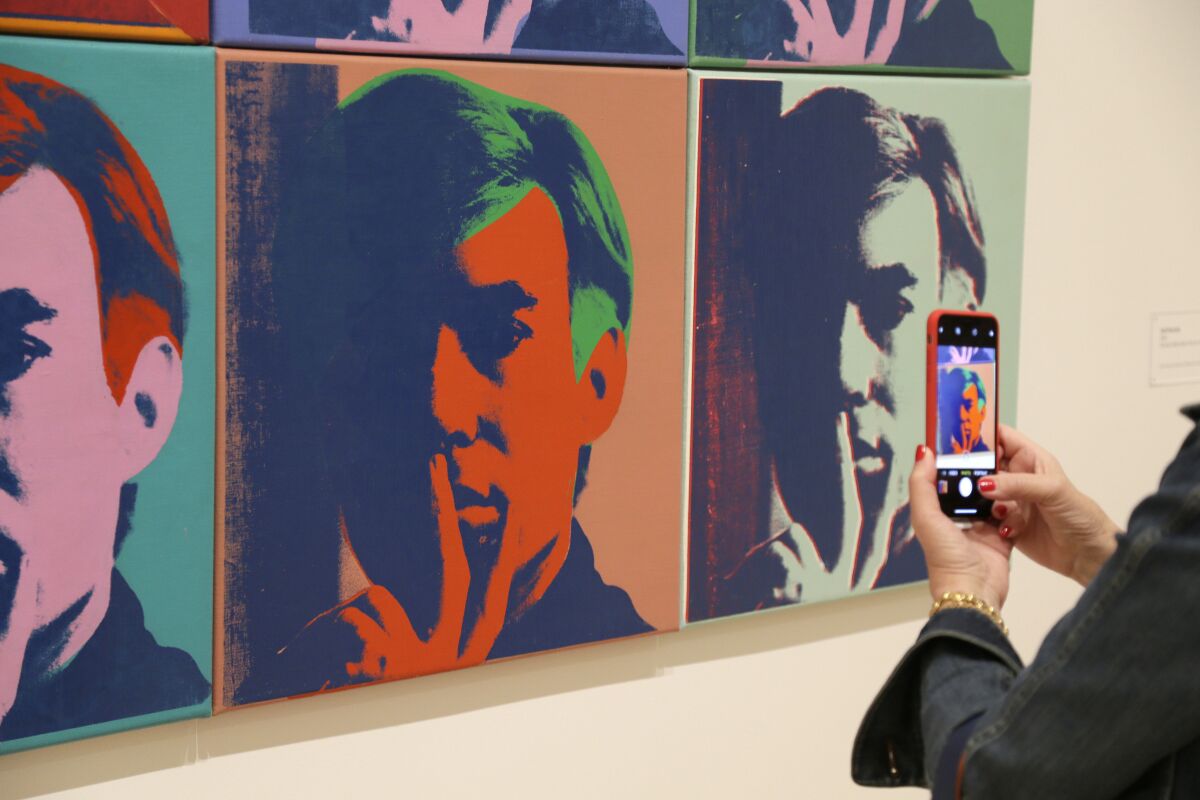 A woman takes a picture of a row of Andy Warhol's silkscreened self-portratis in a museum gallery