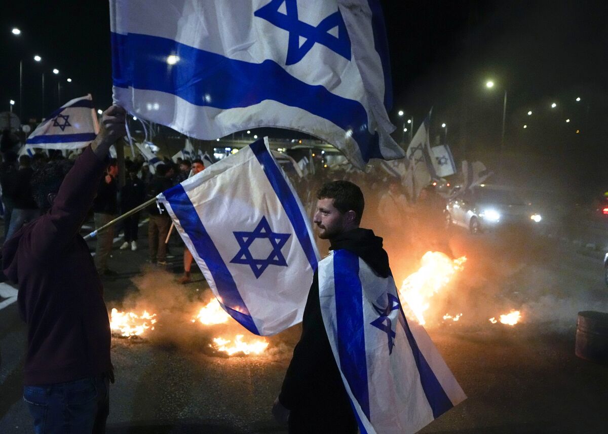 A man wears the Israeli flag as cape during a nighttime demonstration as protesters burn tires nearby.