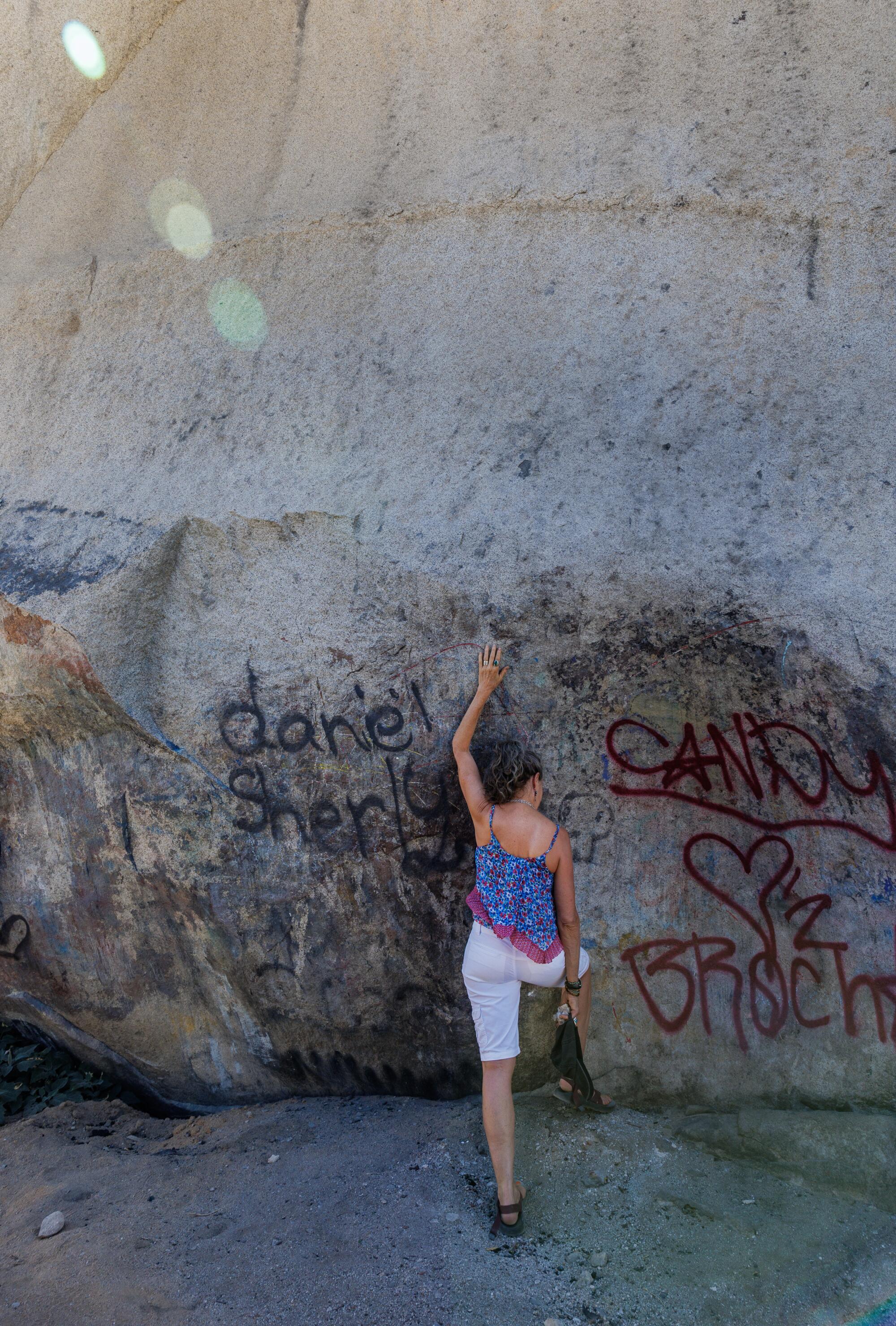 A woman stretches her hand over her head to place it on the surface of Giant Rock, which is scarred by graffiti. 