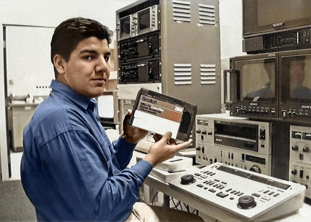 A high school boy holds up a video tape in front of AV equipment 