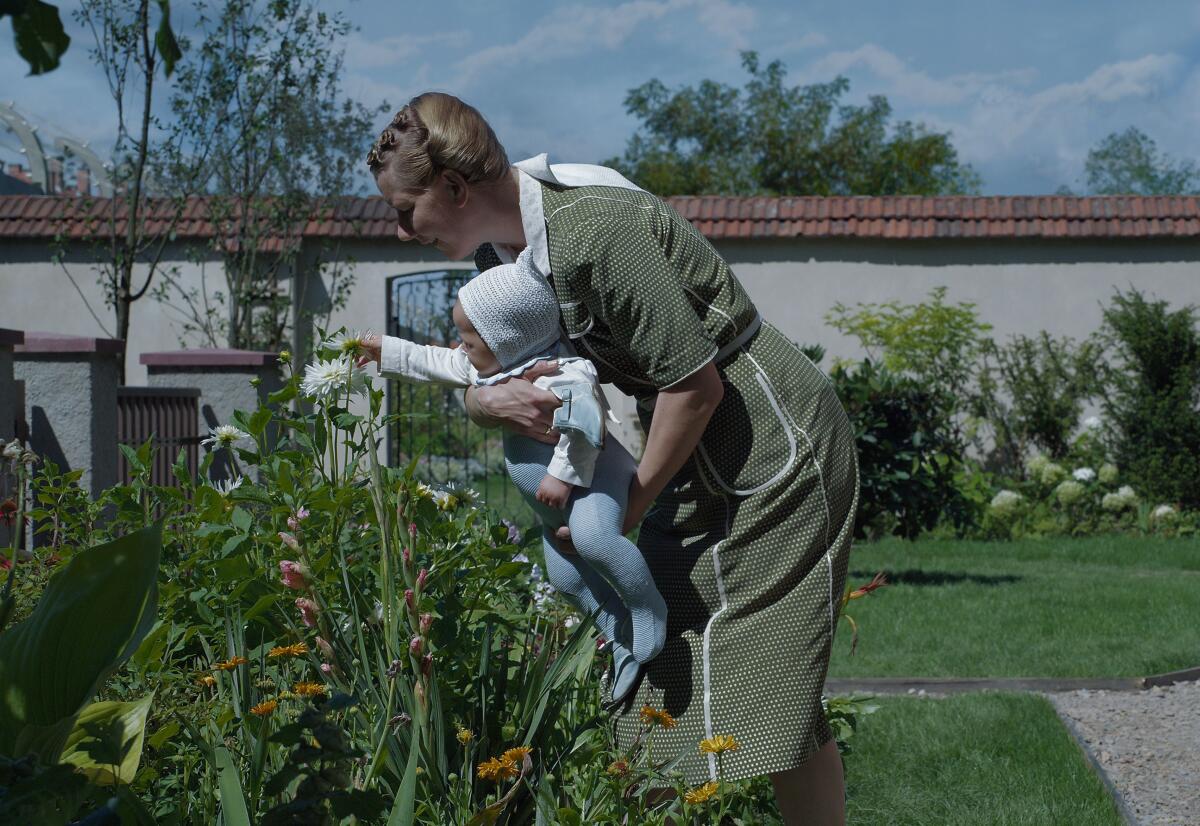 A woman holding a baby bends so the baby can reach out to touch a plant in a garden.