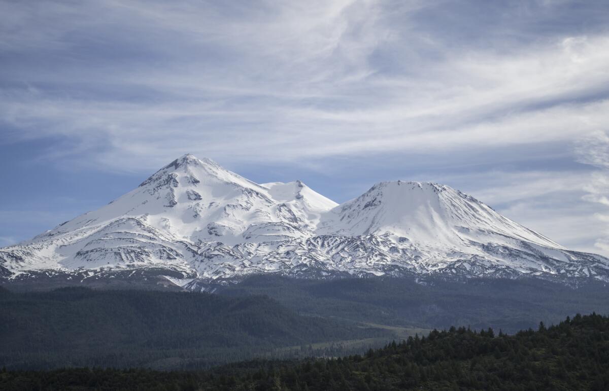 Seen from a distance, mountains are encased in snow, with green, tree-covered hills in the foreground.