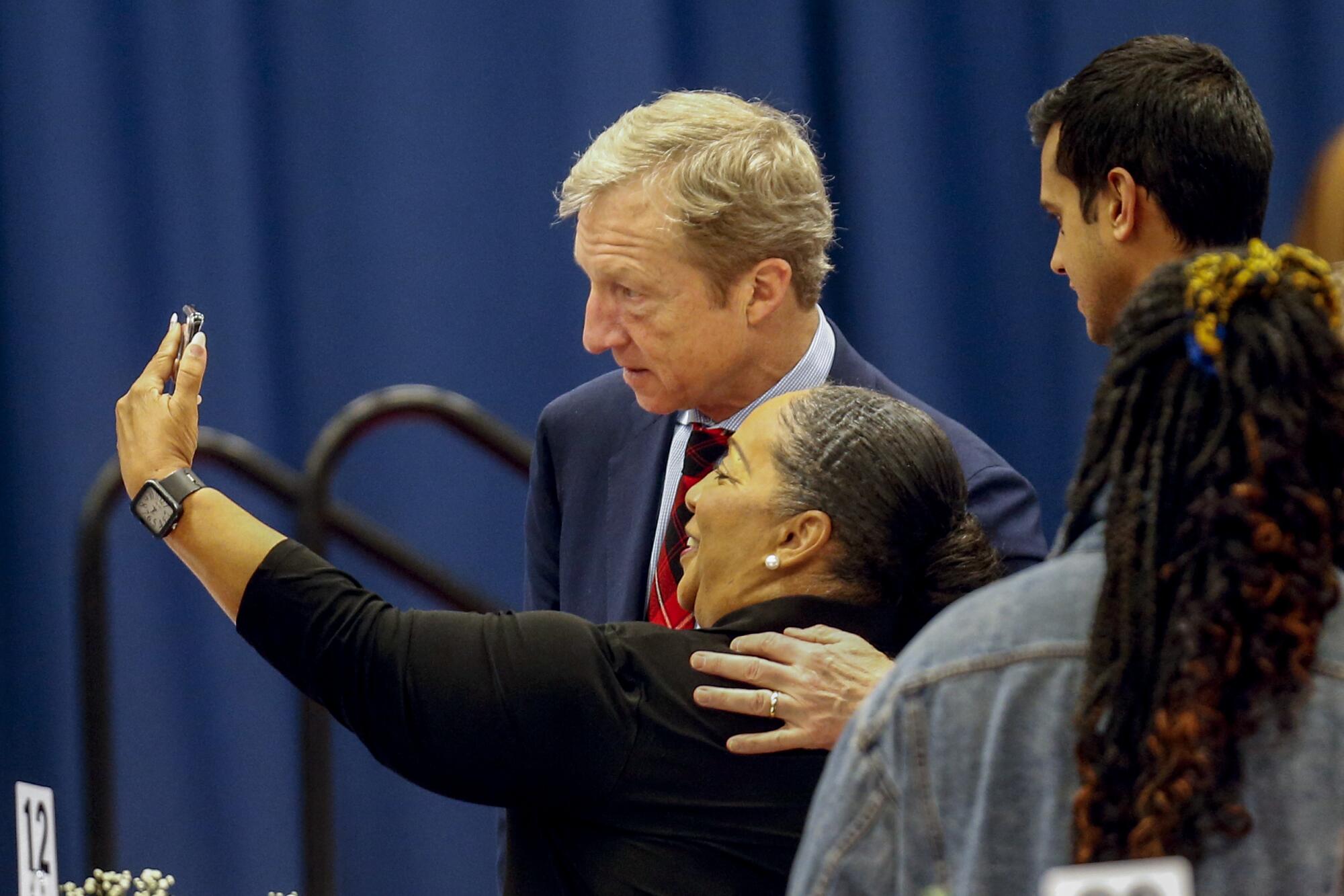 Tom Steyer wearing a dark blue suit and side-hugging a guest as they take a selfie