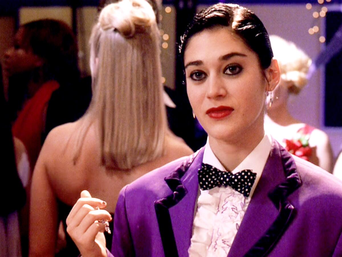 A young woman with slicked-down dark short hair, wearing a purple tuxedo, looks straight ahead