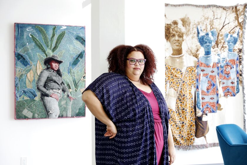 Artist April Bey, in a dotted blue robe, stands before paintings in her studio.