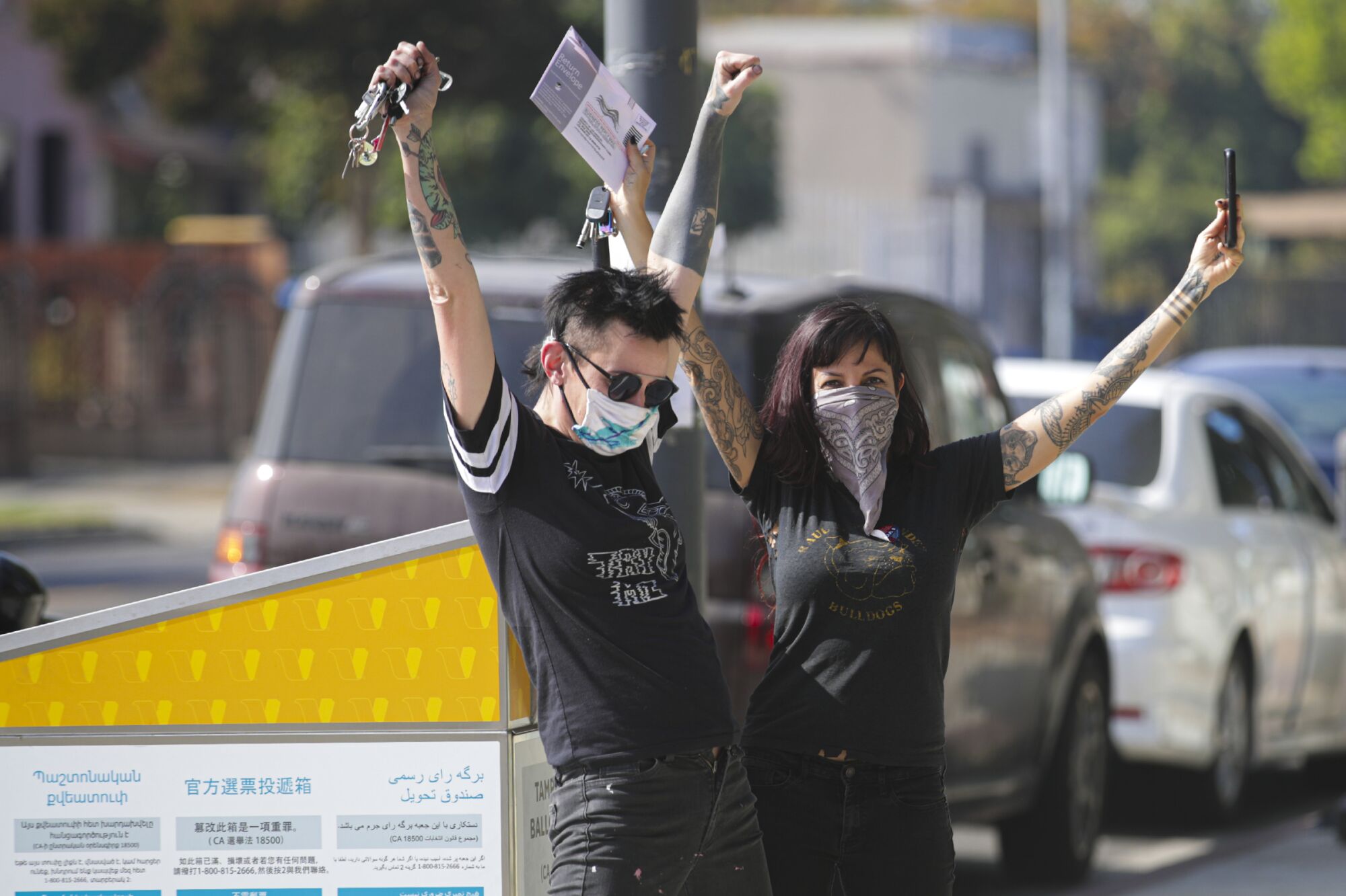 Two young people lift tattooed arms to the sky in front of a ballot box.