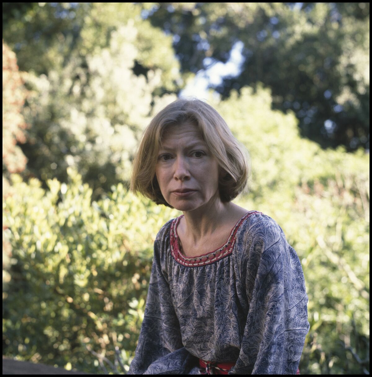 Joan Didion is seen seated in a garden in a loose floral blouse.