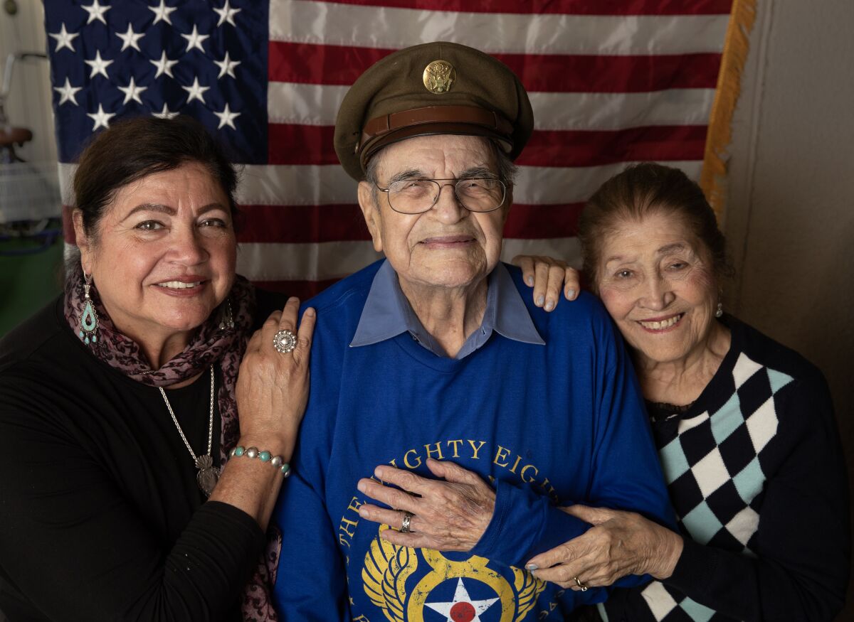 An older man in a military hat holds his hand on his chest while two women flank him in front of a U.S. flag.