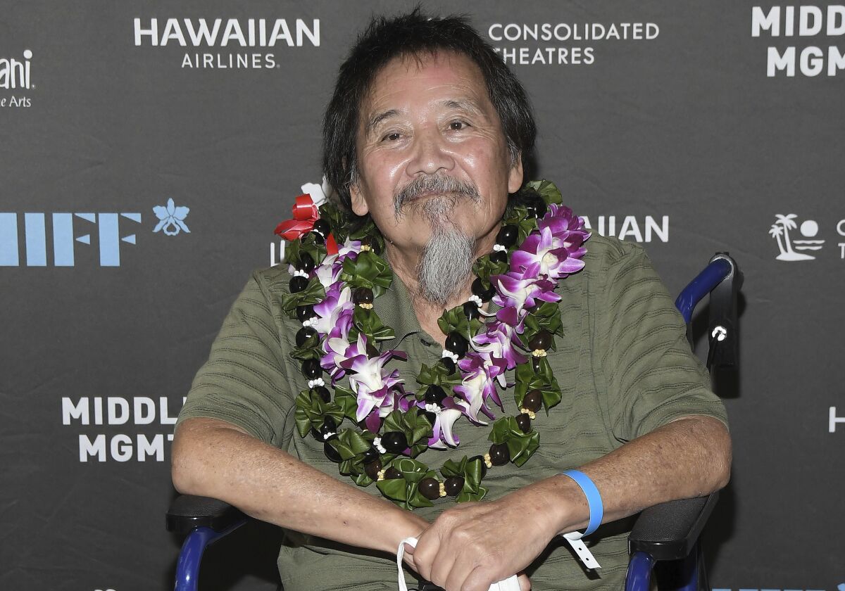 A man in a wheelchair with dark hair and a gray beard wearing a green shirt and multiple leis around his neck