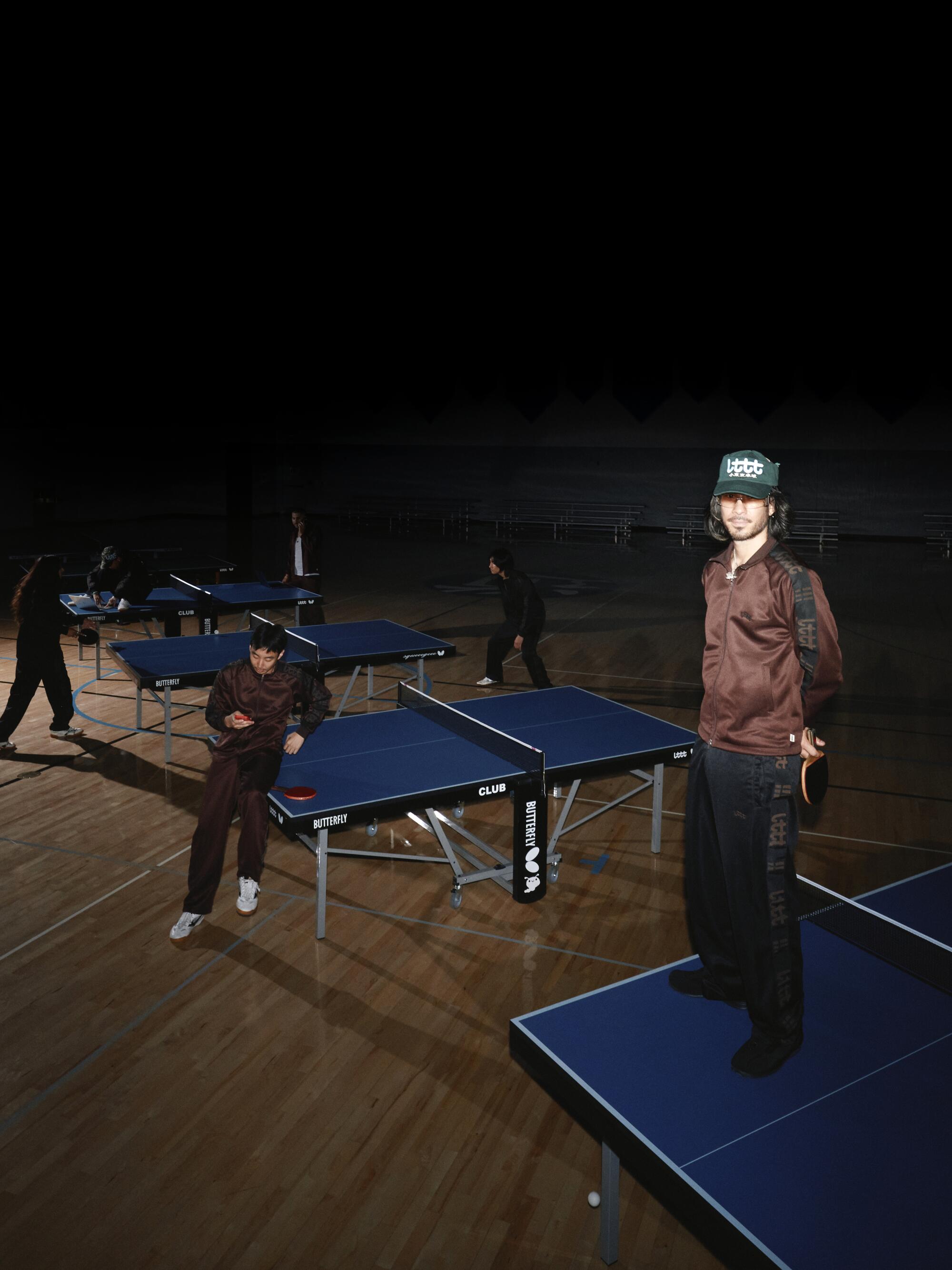 A man stands on a table tennis table, while other people play table tennis in the background.