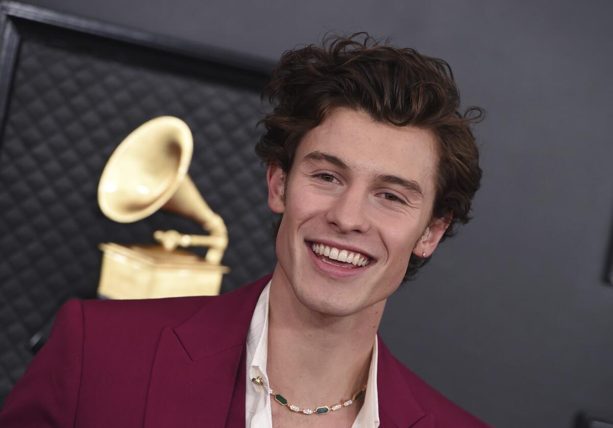 A young man with curly brown hair wears a maroon suit and smiles