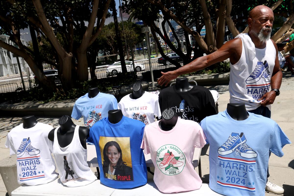 A man shows off an outdoor display of various Kamala Harris and Harris-Walz T-shirts for sale, many of them with Converse sneakers.