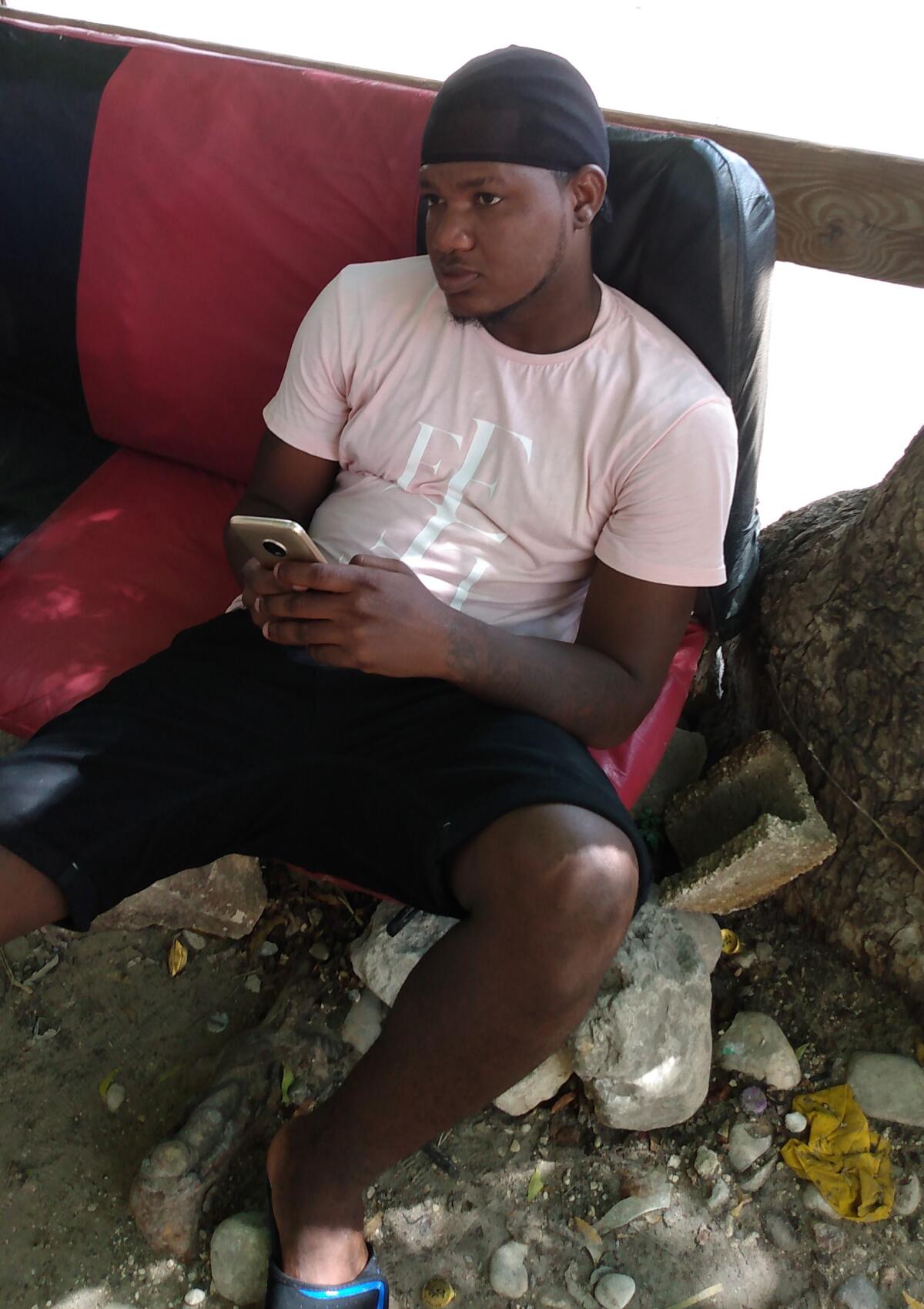 A man in a T-shirt and shorts, holding his cellphone, sits on a bench seat ripped out from a vehicle 