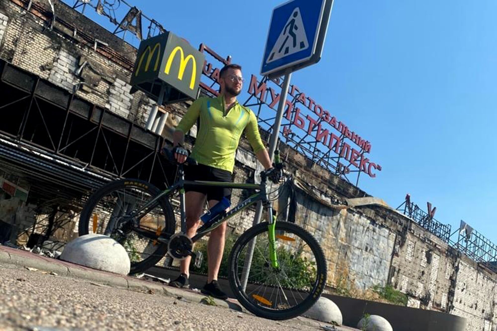 A bearded man in a lime-green shirt and dark shorts holds his bicycle in front of a building with scorched bricks