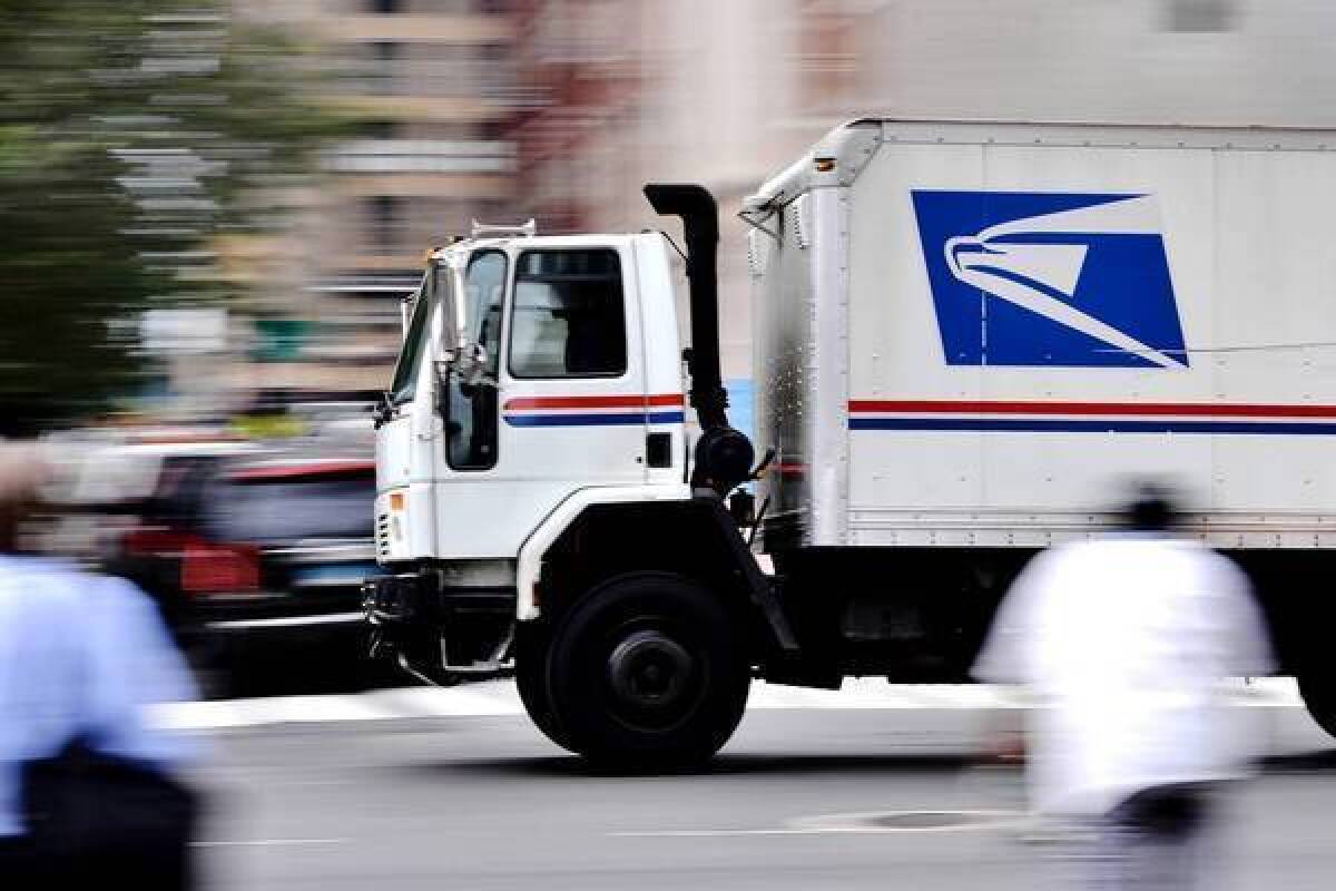 A U.S. Postal Service truck, in focus against a blurred background, drives on a city street