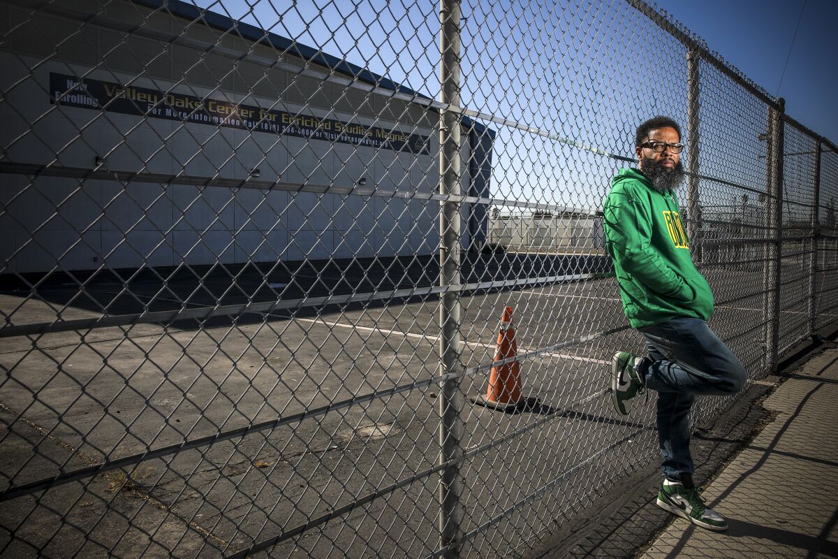 A man with his hands in the pocket of a sweatshirt leans against a school chain link fence.