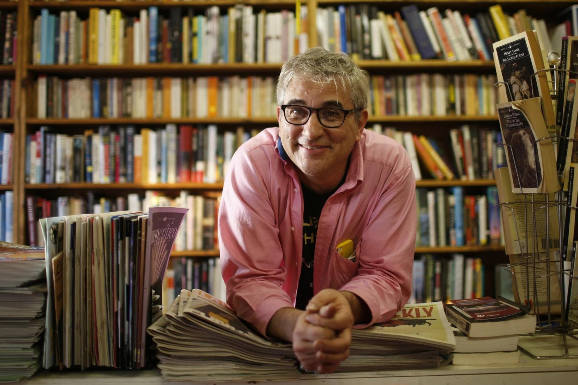 David Kipen leans on a stack of publications on a table with shelves of books in the background.