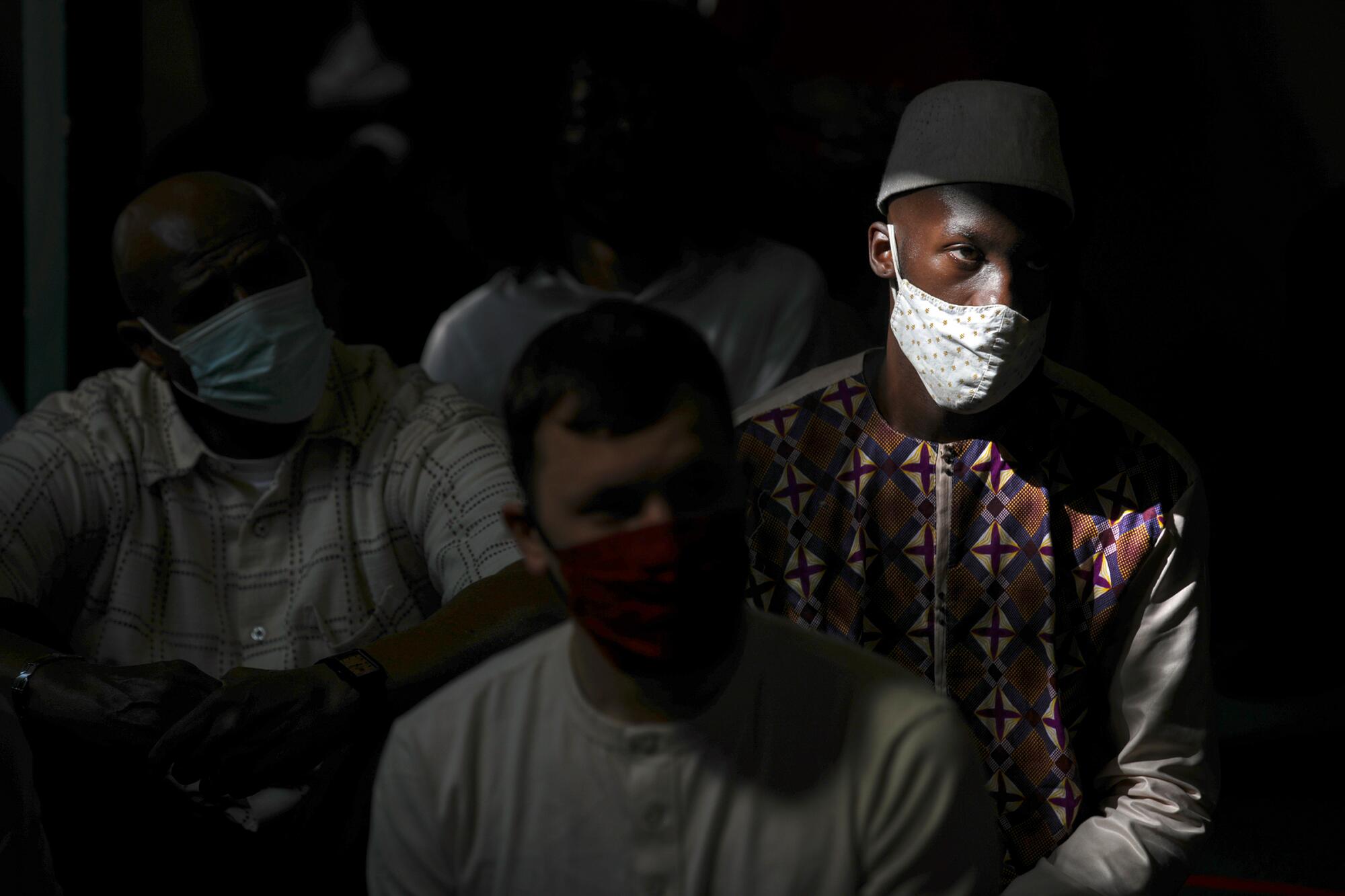 A man wearing a mask and hat sits next to other men, also masked