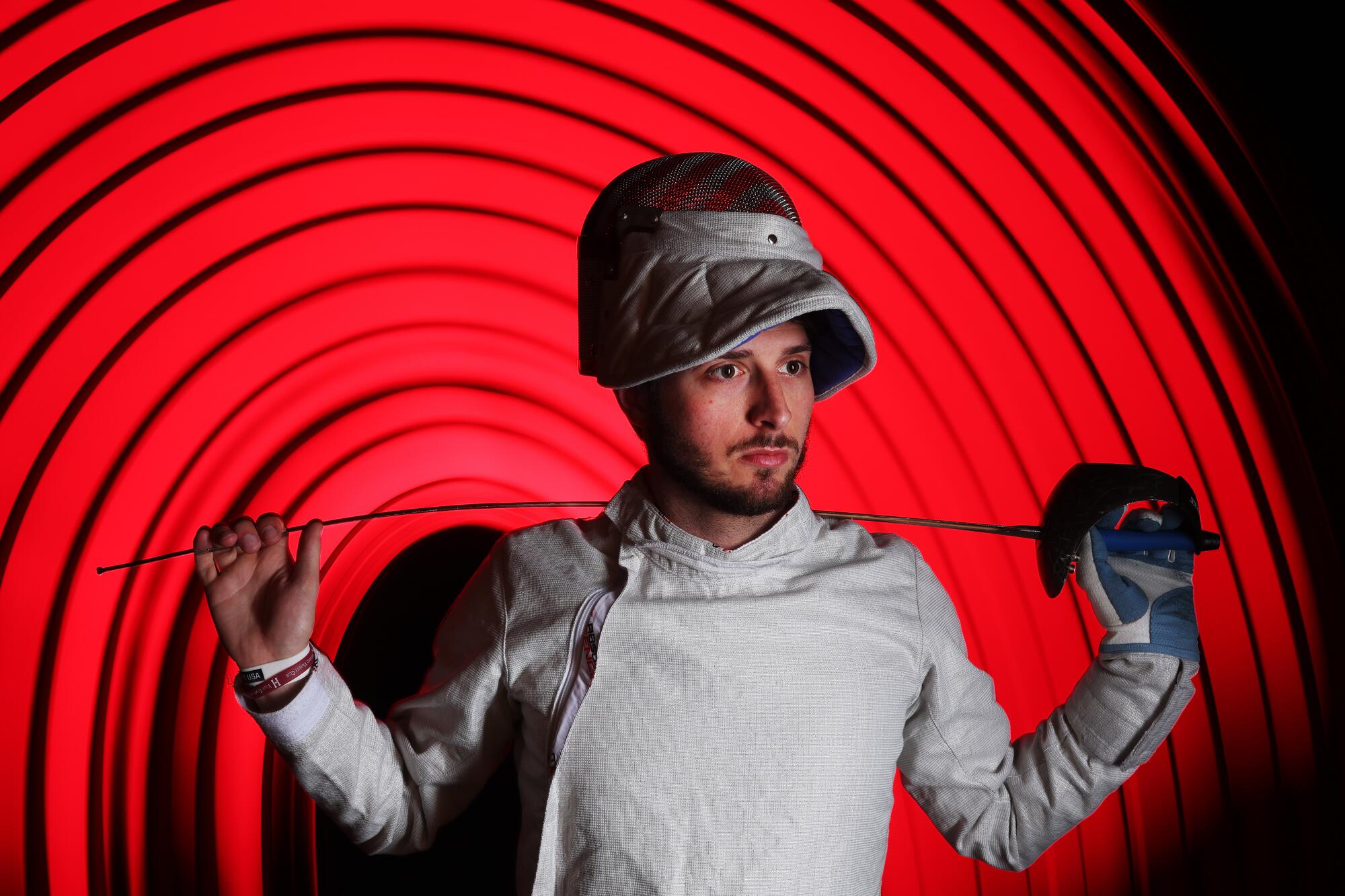 Saber fencer Eli Dershwitz poses in front of a red background.