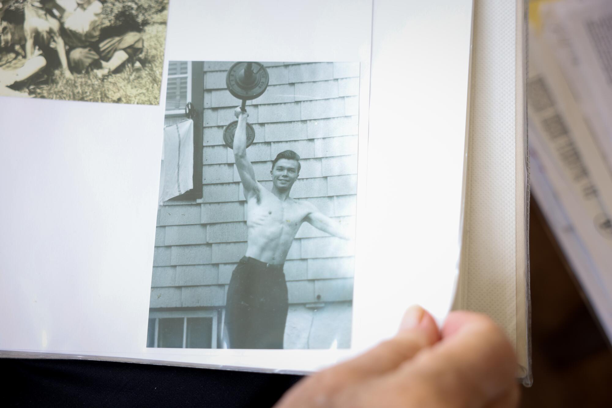 A man holds a page of a photo album with a picture of a man lifting dumbbells with one arm.
