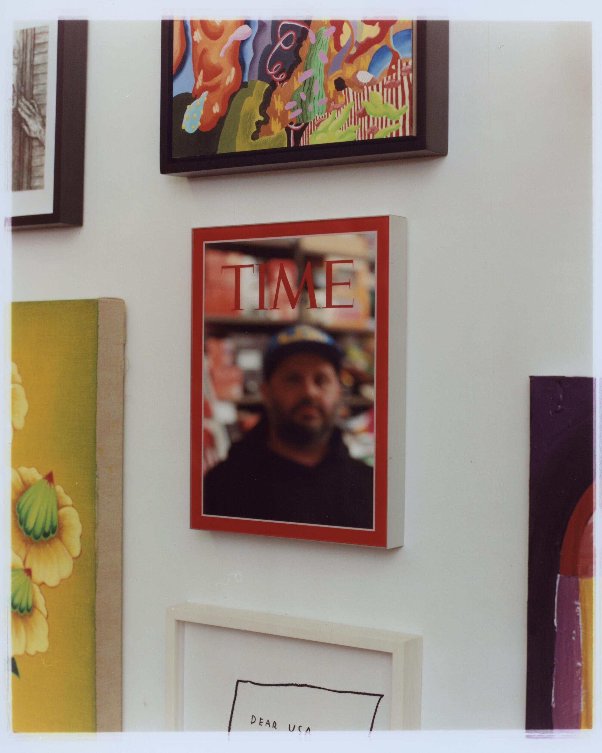 A bearded man in a baseball cap reflected in a mirrored fake Time magazine cover hanging on a wall