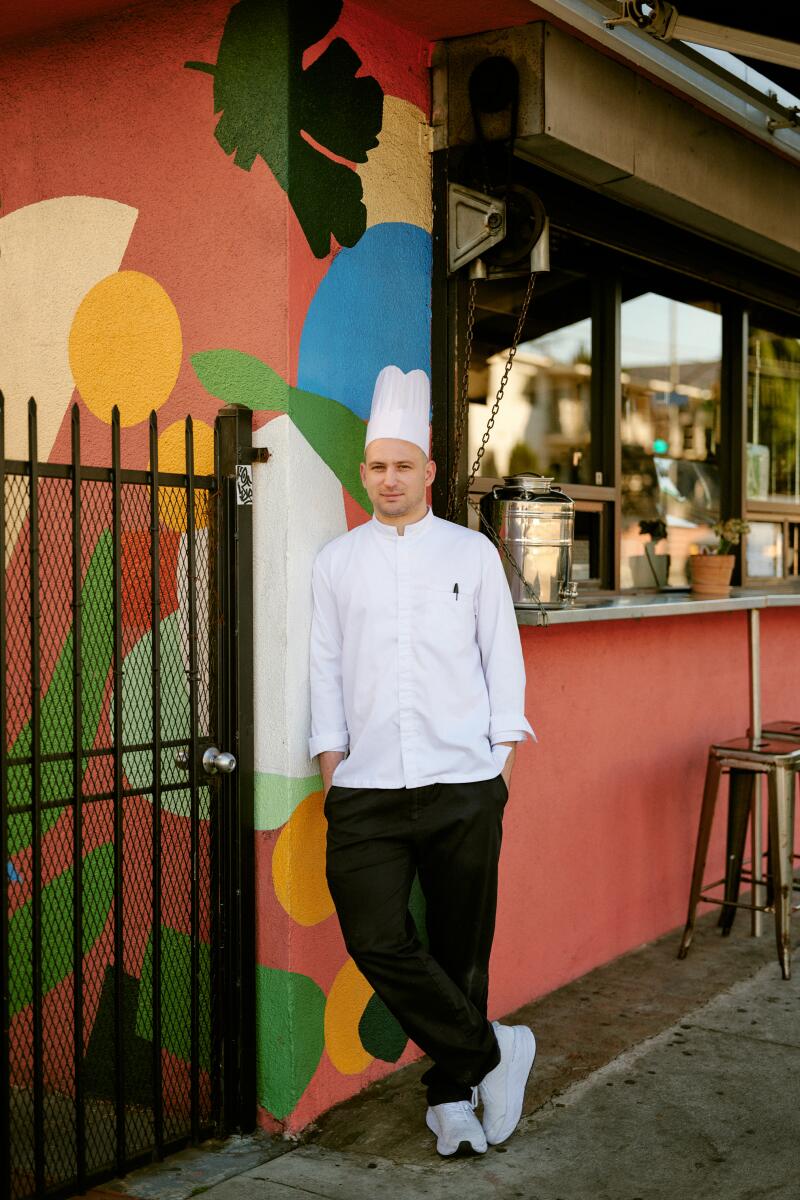 A man in a chef's white hat and jacket leans against a colorfully painted building's wall.
