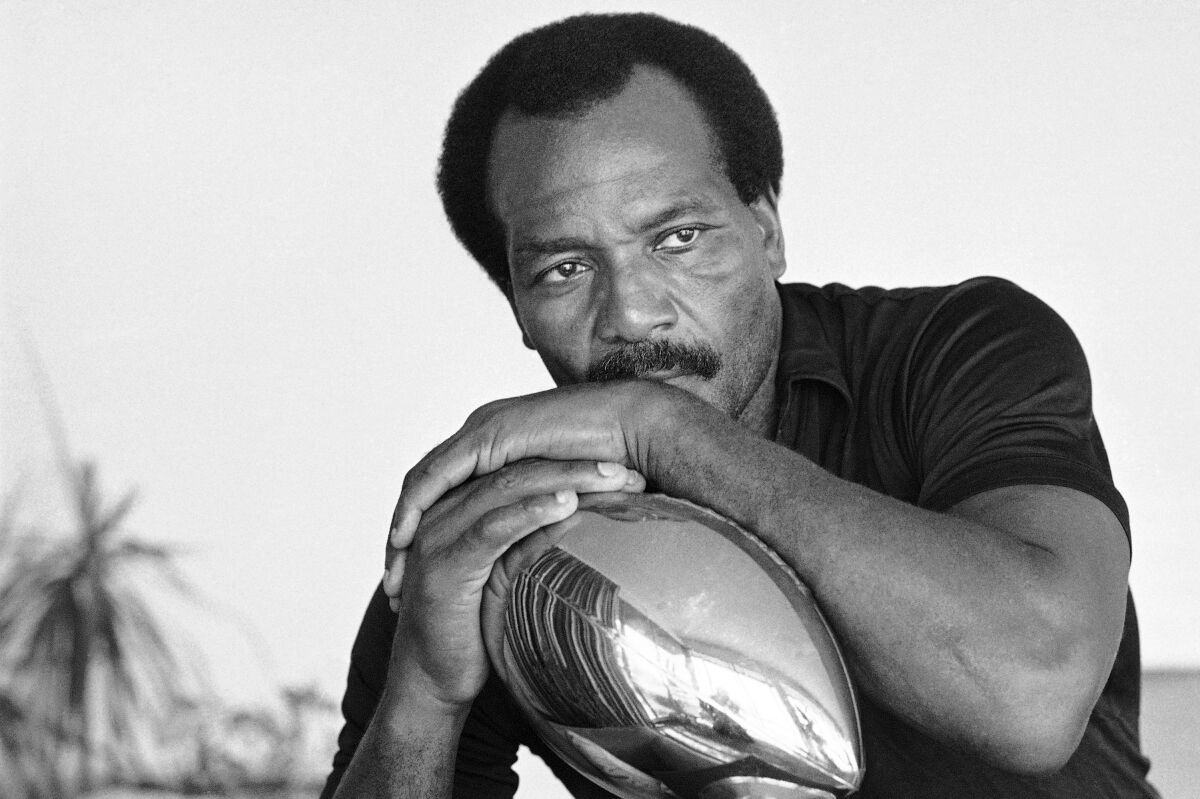 Jim Brown sits in his home while leaning with his hands folded over a football-shaped trophy.