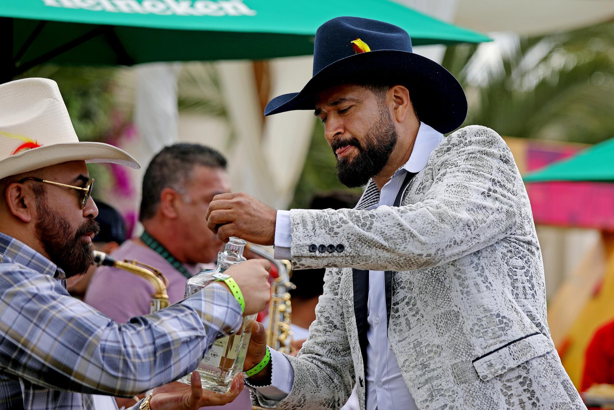 A bearded man wearing a jacket and cowboy hat hands other cowboys shots of tequila. 