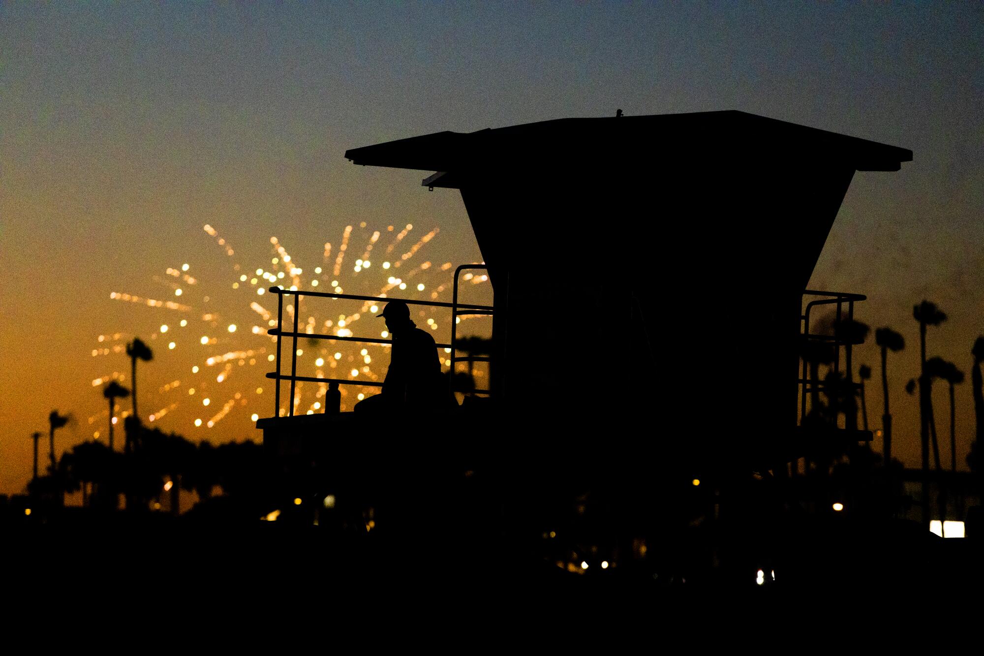 A silhouette of a person and lifeguard tower with orange sparks from fireworks against an evening sky 