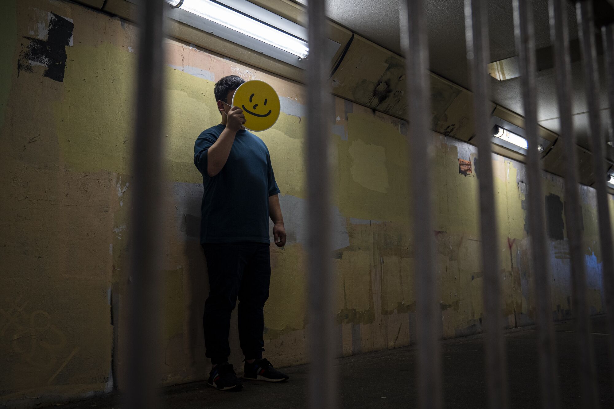 A man holds a yellow smiley face sign in front of his face in a dimly lit hallway, seen through metal bars