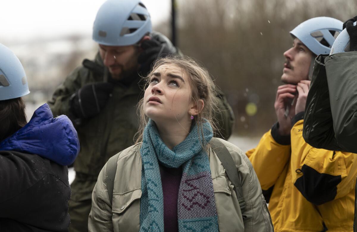 A woman in a jacket and scarf, surrounded by men in blue bike helmets, looks up into the sky.