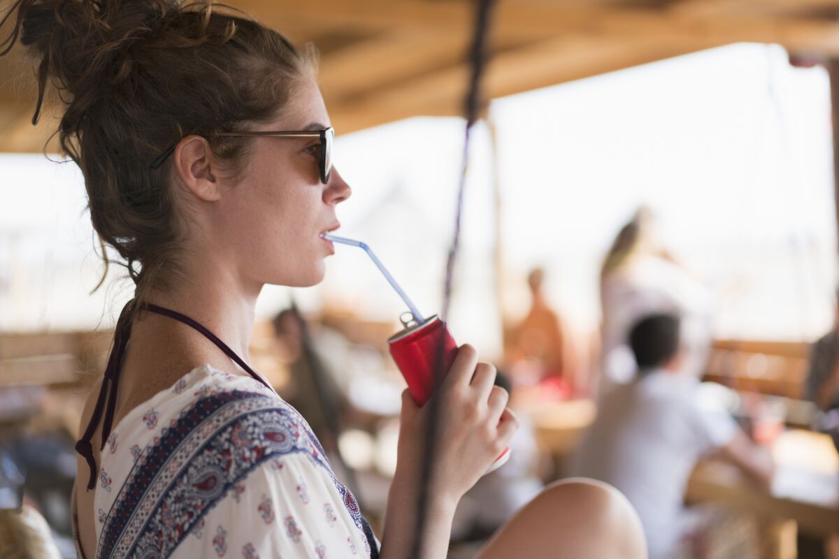 A young woman at the beach sits at a bar, drinking something out of a can.