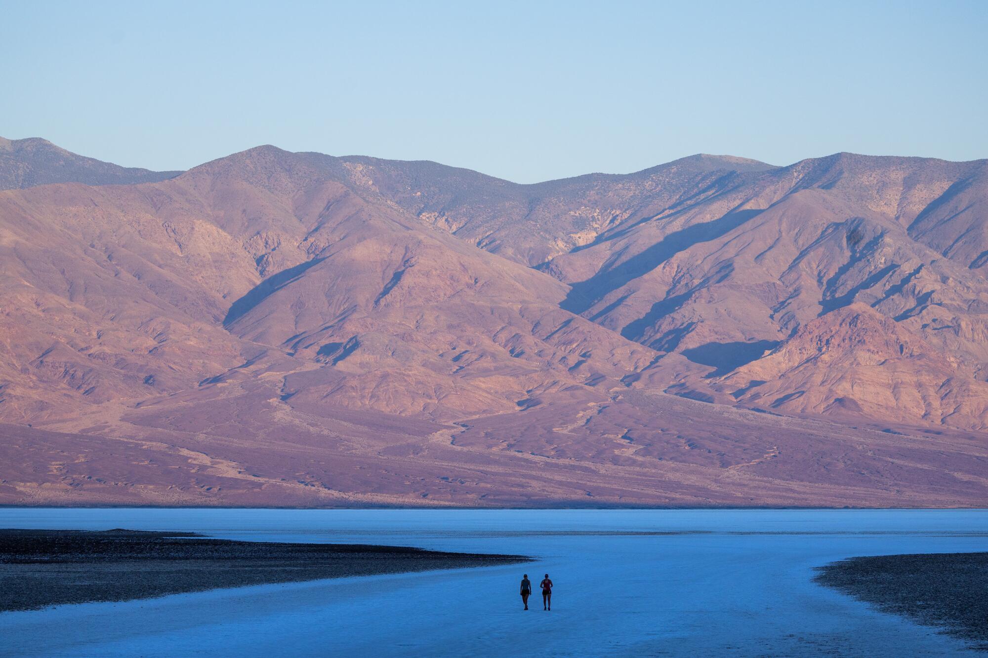 Two figures walk on a salt flat as desert mountains rise in the background.