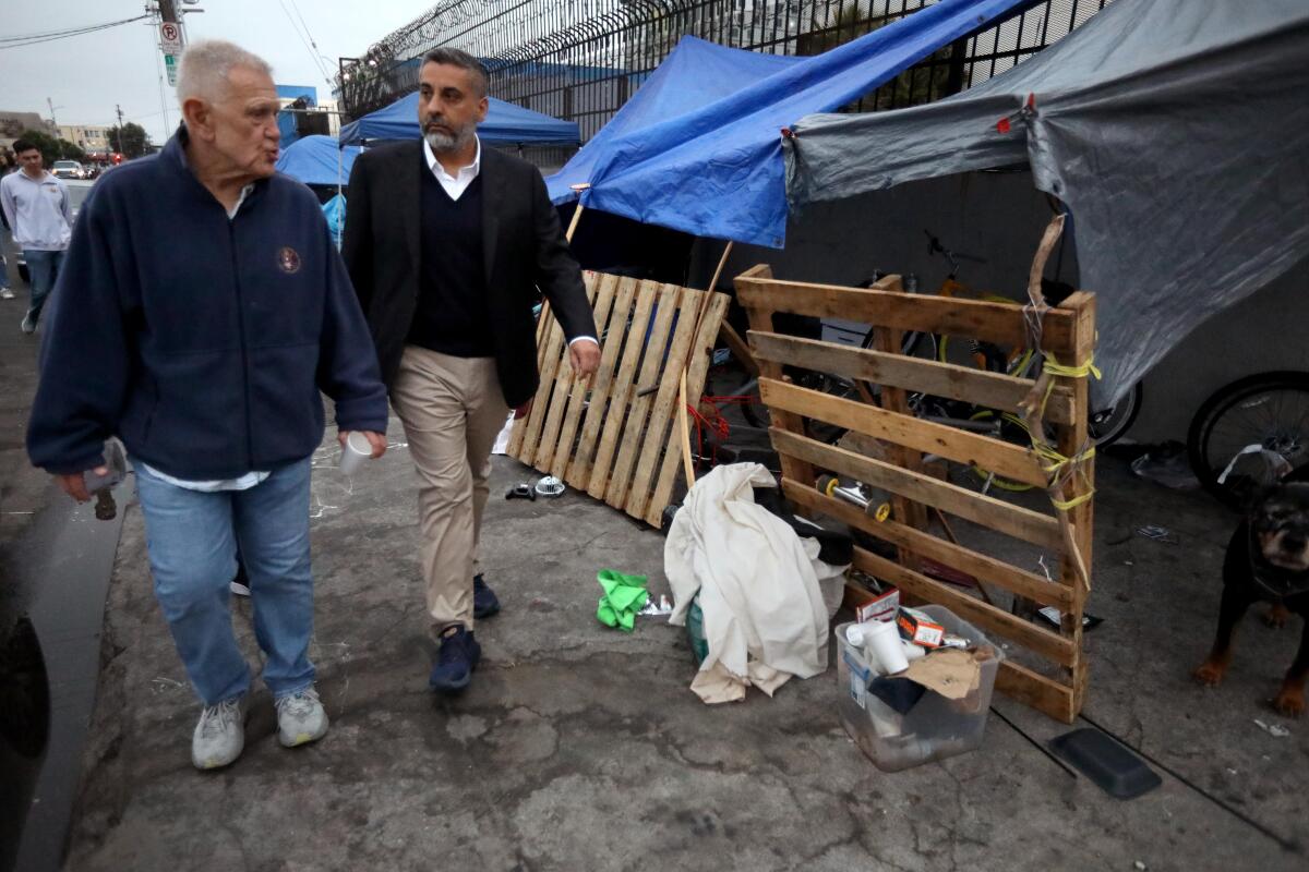 Two men walk past tents with pallets on one side on a sidewalk.