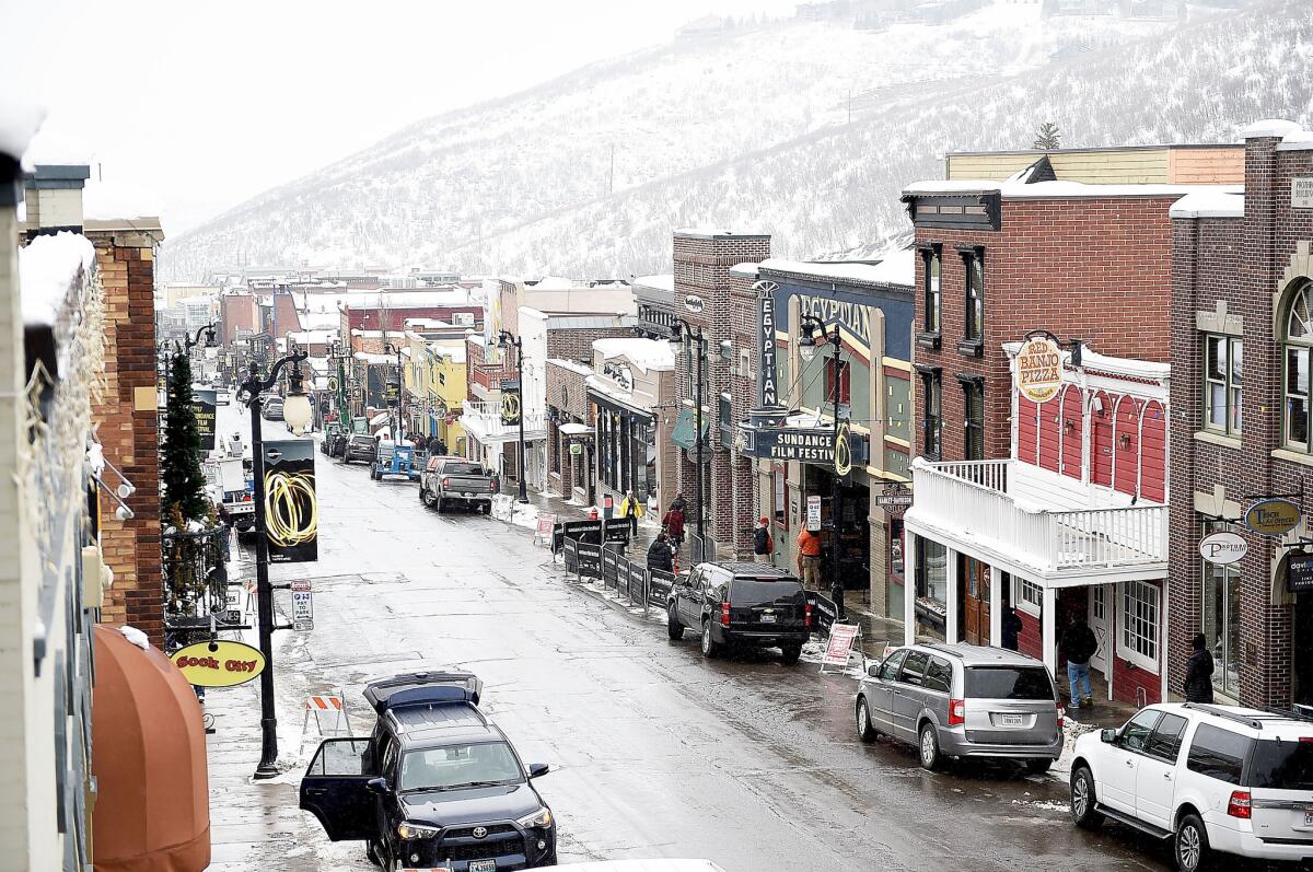 A street of store fronts and parked cars with snow covered hills in the background.