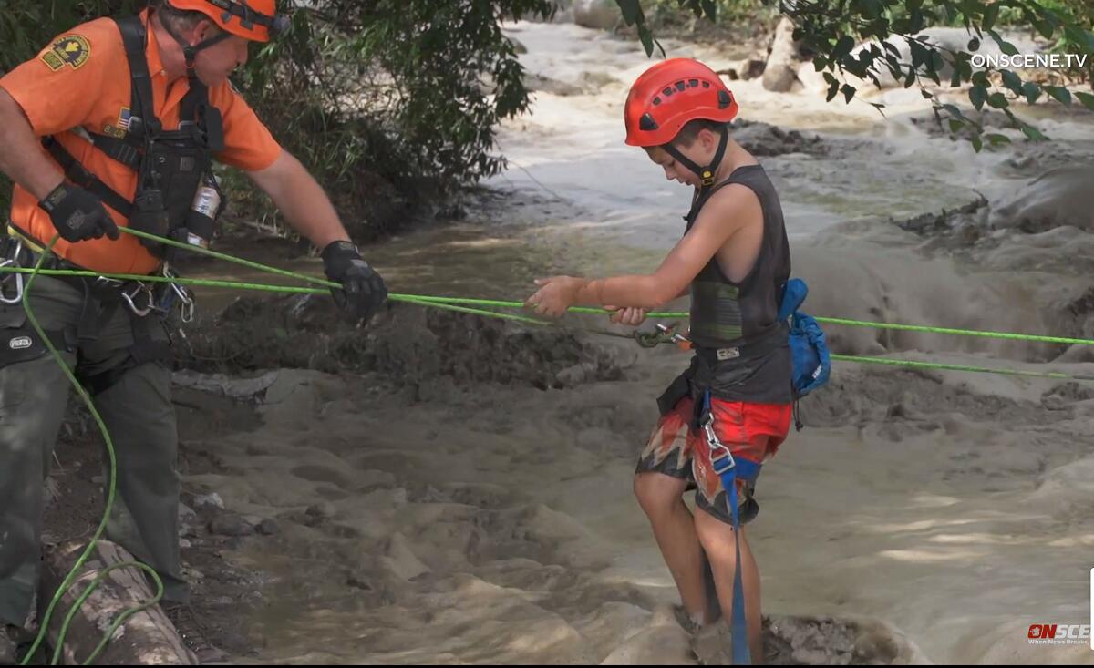 An emergency worker helps a youth cross floodwaters while holding two rope lines.