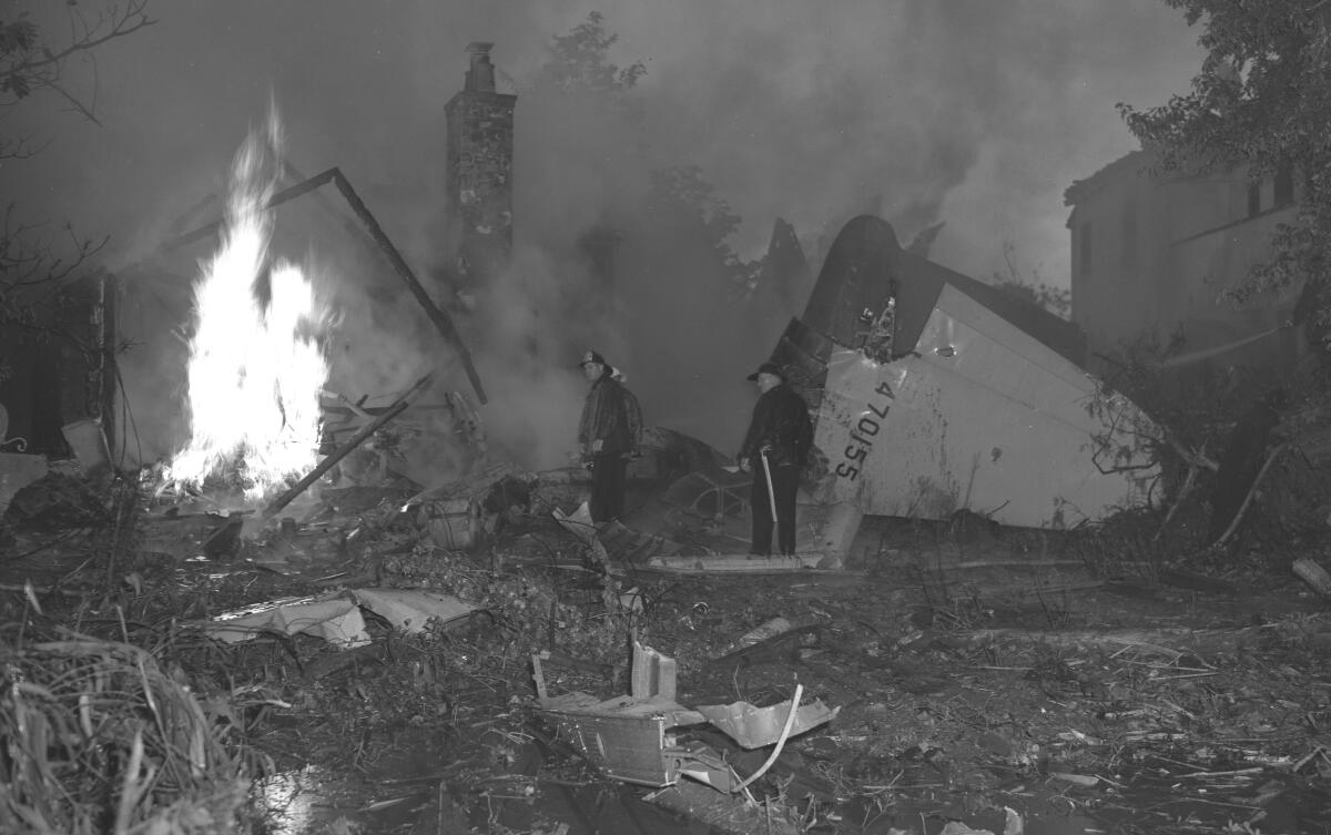 In the dark and haze two men in uniform stand between an oversized chunk of plane and leaping flames.