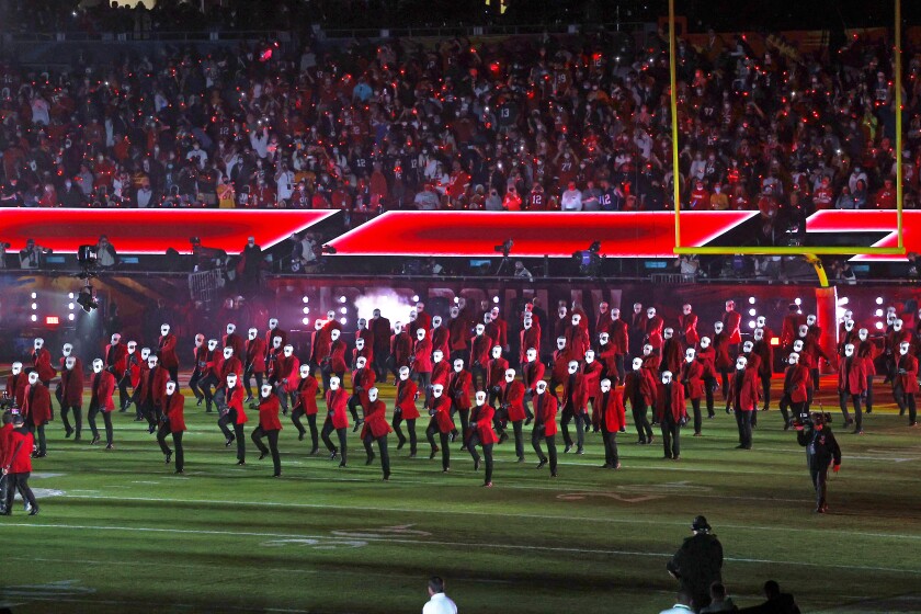Performers in bright red jackets and white face masks march into a football field for a halftime show