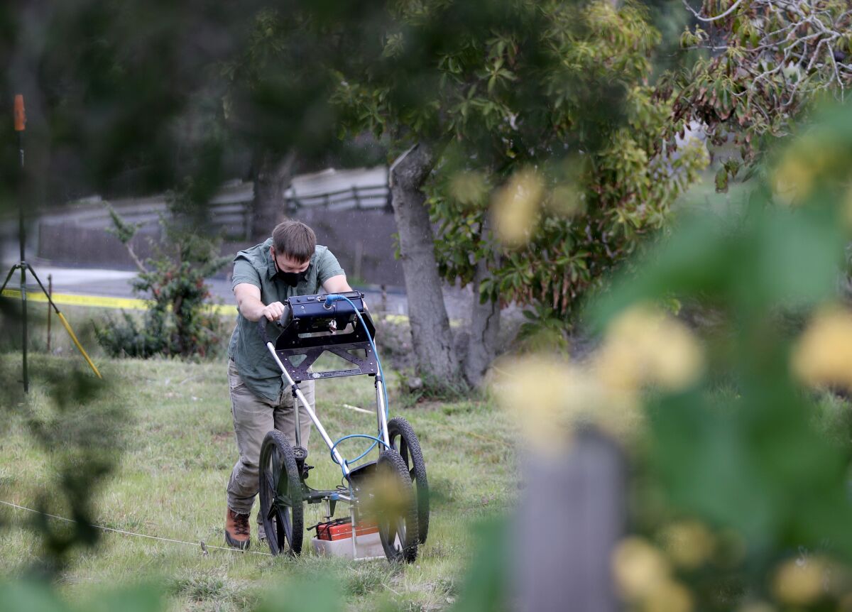 A man in a mask pushes a wheeled device among trees in a yard.