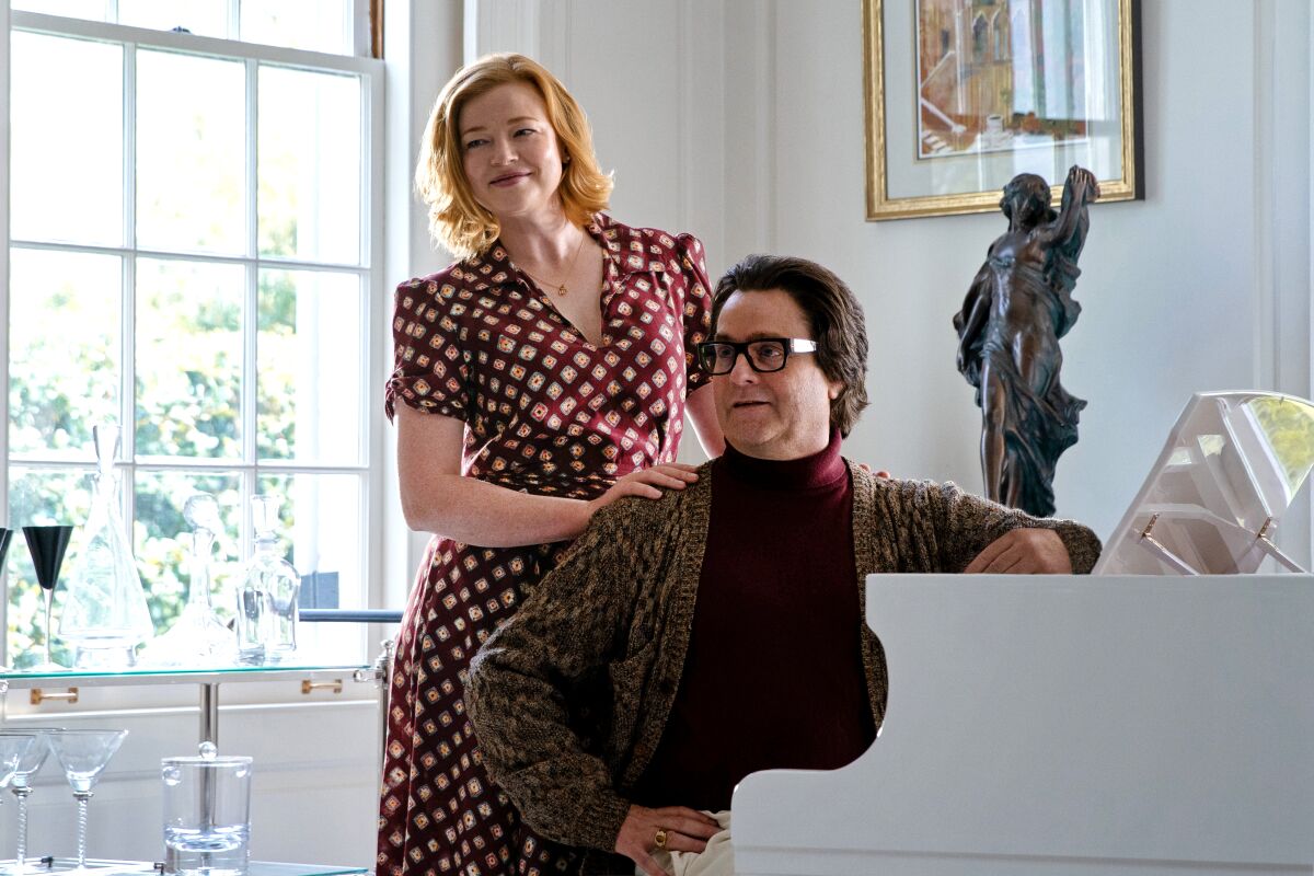 A smiling woman stands behind a man seated at a white piano