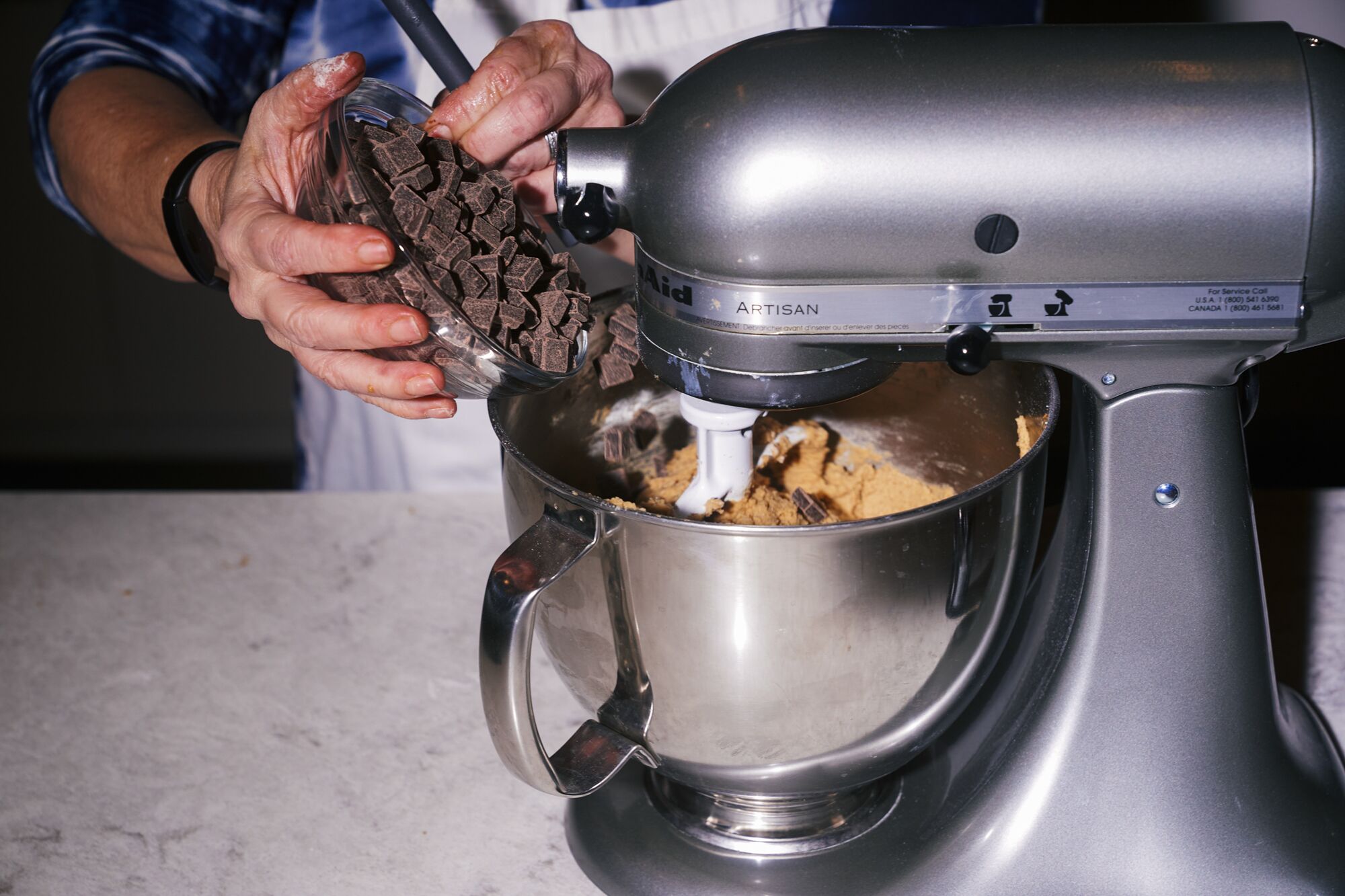 Hands pour a bowl of chocolate chunks into a stand mixer.