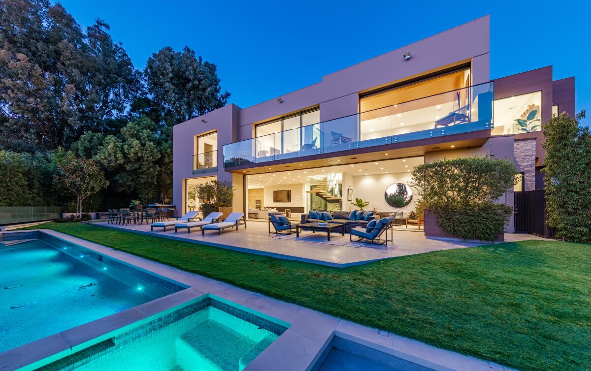A backyard pool and patio and modern multistory house are shown in the evening with lights glowing.