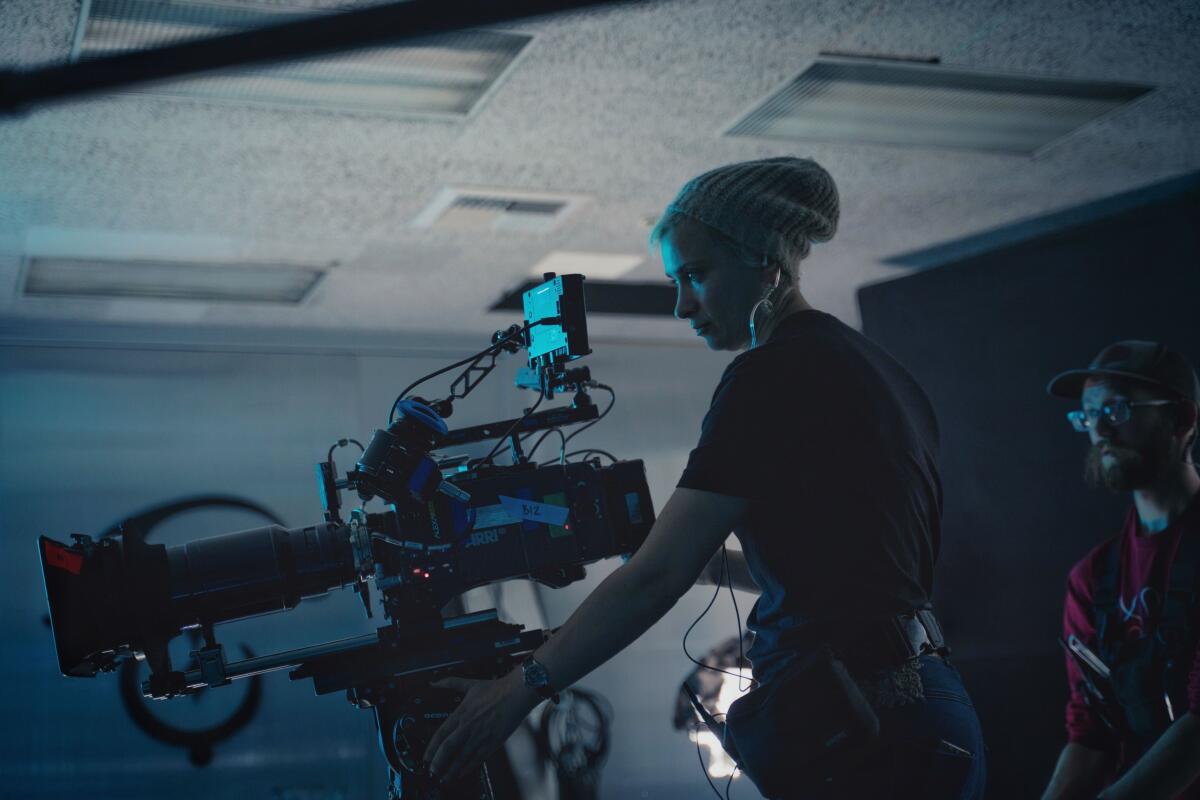 Cinematographer Halyna Hutchins stands next to a film camera in a dark room while a man looks on.