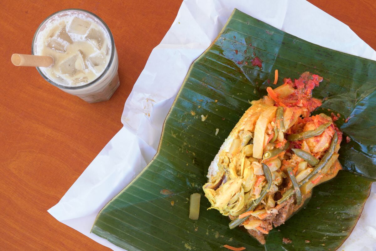An iced drink on a table next to banana leaf-wrapped nasi bungkus with chicken curry and beef rendang